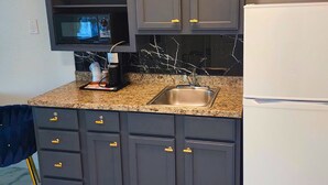 A kitchen with dark cabinets, a granite countertop, a stainless steel sink, a built-in microwave, and a white refrigerator. - The Enchanted Suites & Residence  (Roswell)