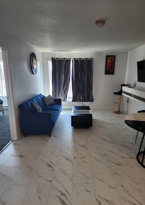 A modern living room with a blue sofa, a black ottoman, a marble floor, a TV, and a dining area with a table and chairs. - The Enchanted Suites & Residence  (Roswell)