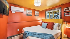 A hotel room with a bed, an air conditioning unit, a window with a view, and artwork on the walls.