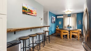 A kitchen with a wooden bar counter, stools, a dining table with chairs, and a refrigerator.