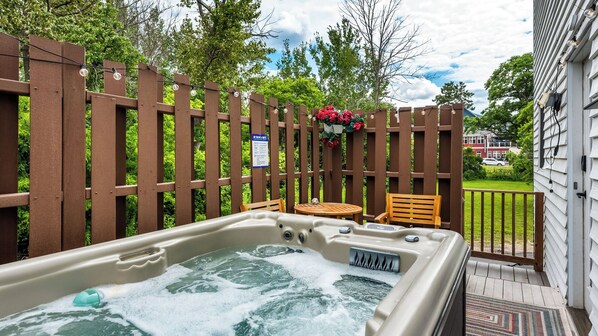 A hot tub with a wooden deck and chairs, surrounded by a wooden fence and greenery.