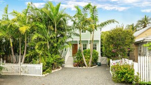 A house with a front porch, surrounded by palm trees and a white picket fence.