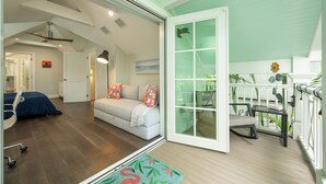 A spacious living room with a sofa, a desk, and a chair. There is a sliding glass door leading to an outdoor area with plants.