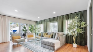 A modern living room with a grey sofa, a glass coffee table, and a patterned rug. There are two potted plants and a large window with curtains. - Modern Lush Green Escape (Fort Lauderdale)