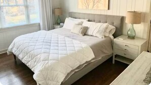 Ein ordentlich gemachtes Bett mit weißer gesteppter Decke, ein Nachttisch mit Lampe und ein an der Wand hängendes Kunstwerk darüber.