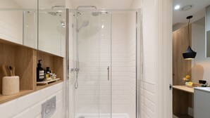 A modern bathroom with a glass-enclosed shower, white tiled walls, a wooden vanity, and a pendant light. - Cozy Suite in Sloane Square (London)