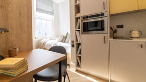 A compact bedroom with a bed, bedside table, and a window with blinds. Adjacent is a kitchenette with a microwave, oven, and a coffee maker. - Cozy Suite in Sloane Square (London)
