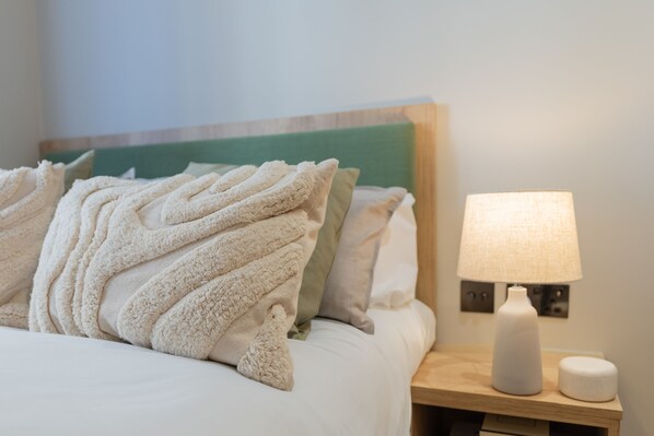 A neatly made bed with a fluffy white blanket and pillows, a bedside table with a lamp and a small container, and a green headboard.