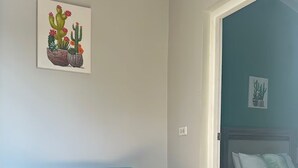Een kamer met een blauwgroene bank, twee oranje kussens, een bed met witte beddengoed en een wand met een kunstwerk van een cactus.