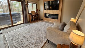 A cozy living room with a large TV, fireplace, and a view of the outdoors. - Hawkins Hideout  Cabin, Starlink Wi-Fi, Hot Tub! (Reed City)