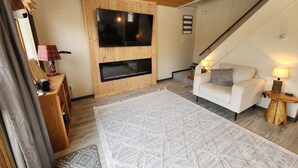 A living room with a wooden ceiling, a flat-screen TV mounted on a wooden wall, a white sofa, two side tables with lamps, and a patterned rug. - Hawkins Hideout  Cabin, Starlink Wi-Fi, Hot Tub! (Reed City)