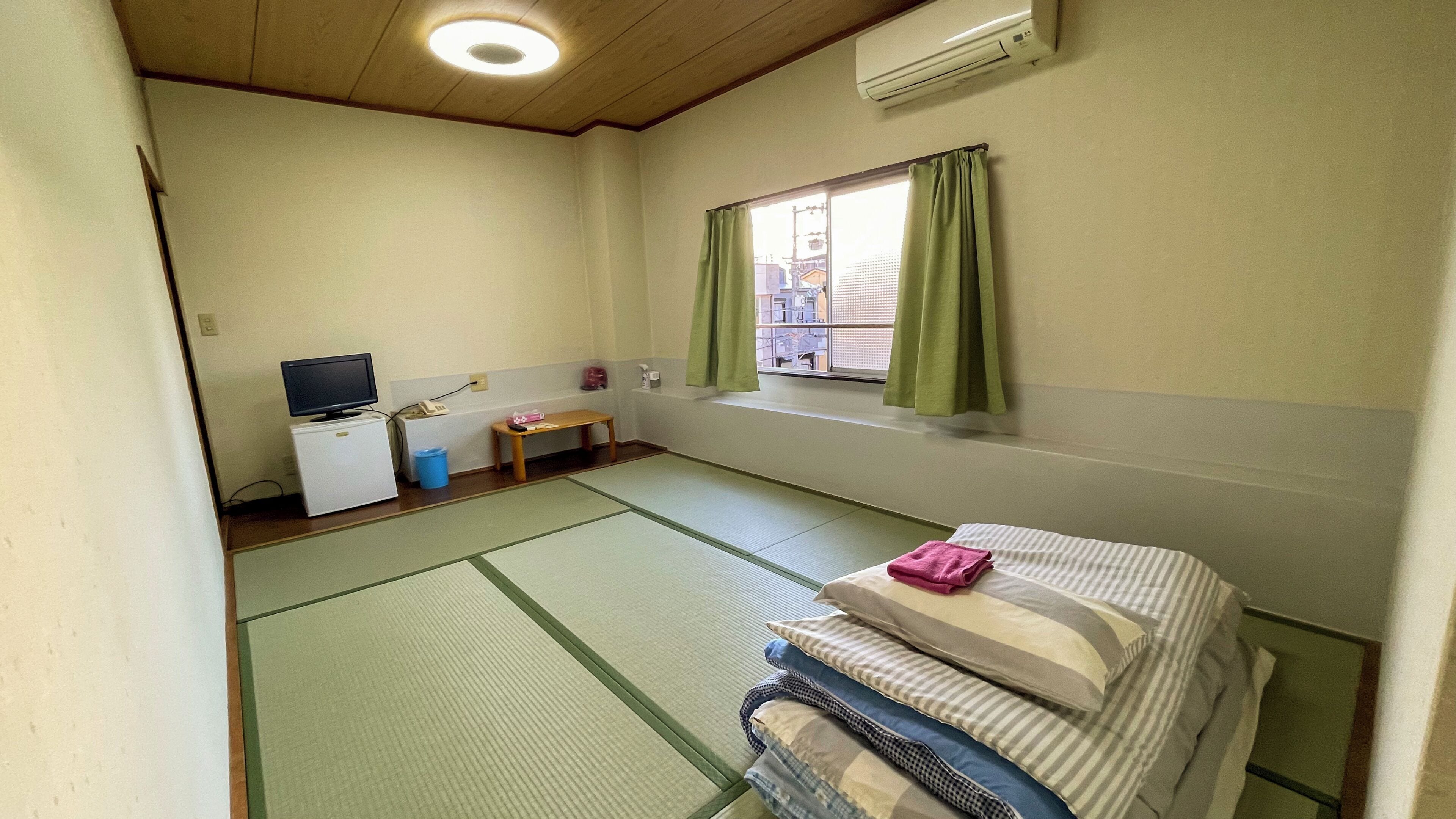 A room with tatami flooring, a bed with pillows, a small table with a TV, and a window with curtains.