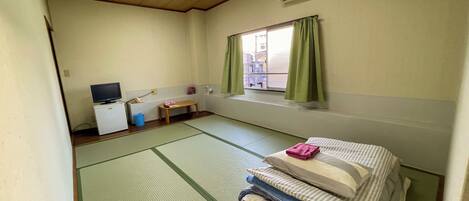 A room with tatami flooring, a bed with pillows, a small table with a TV, and a window with curtains.