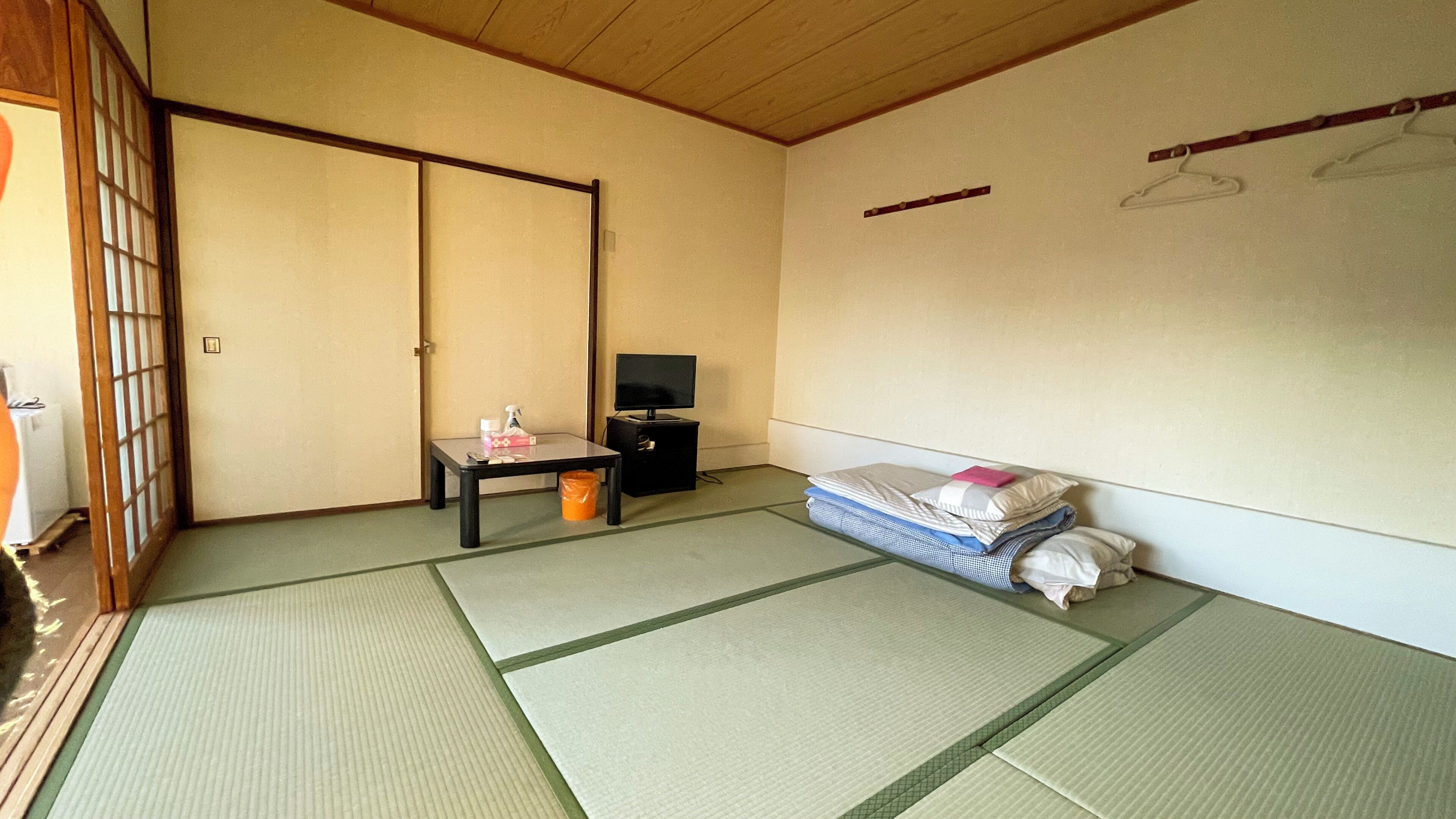 Une pièce de style japonais traditionnel, avec un sol recouvert de tatamis, une porte coulissante, une petite table avec un téléviseur et un lit avec des oreillers.