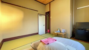 A hotel room with tatami flooring, a bed with a pink towel, a television, and a small table with toiletries. - Nishiki Guest House (Kadoma)