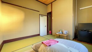A hotel room with tatami flooring, a bed with a pink towel, a television, and a small table with toiletries. - Nishiki Guest House (Kadoma)