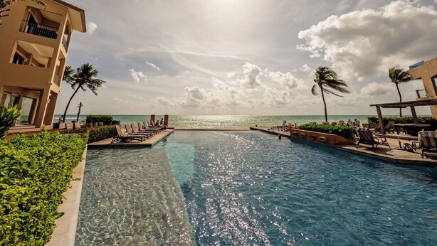 A clear pool with a view of the ocean, surrounded by lounge chairs and palm trees. - Ocean Elegance
Contemporary Beachfront Escape at El Faro Resort Living (Playa del Carmen)