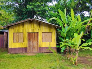 A small, yellow wooden structure with two doors and a single window, surrounded by greenery. - Bamboo cabin with 2 double beds (Catemaco)