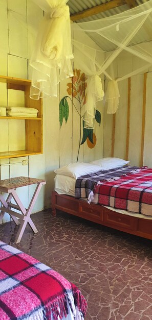 A room with a bed, a wooden shelf, a small table, and a wall with a painted leaf design.