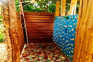 Un petit espace de douche extérieur, avec un sol carrelé, des murs en bambou et un mur bleu percé de trous.