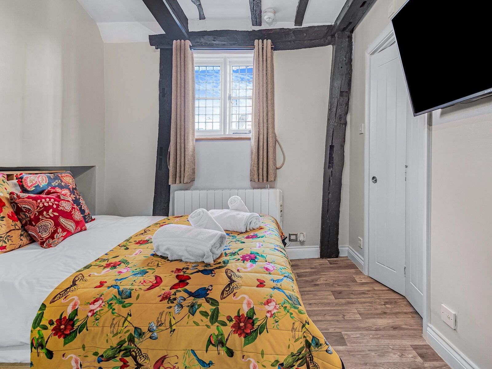 A bedroom with a bed, floral bedspread, two rolled towels, a window with curtains, a radiator, and a television on the wall.
