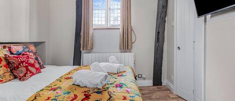A bedroom with a bed, floral bedspread, two rolled towels, a window with curtains, a radiator, and a television on the wall.