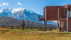 Un edificio moderno con balcón, una montaña al fondo y un área exterior vallada con sillas y una parrilla.