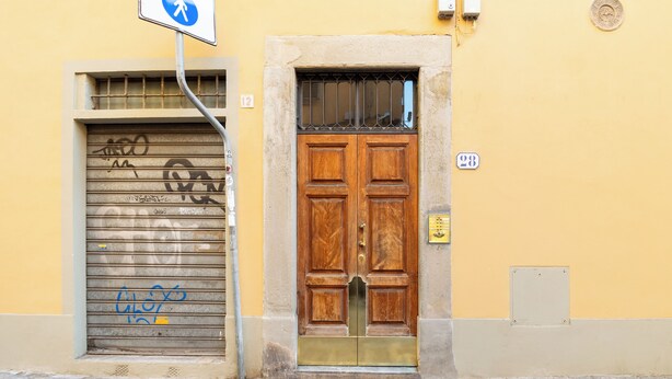 A closed wooden door with a metal bar above it, a metal shutter door with graffiti, and a building number sign. - Via Laura - cozy studio (Florence)