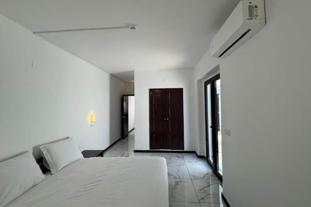 A hotel room with a bed, a nightstand, a wall-mounted light, a door, and an air conditioning unit.