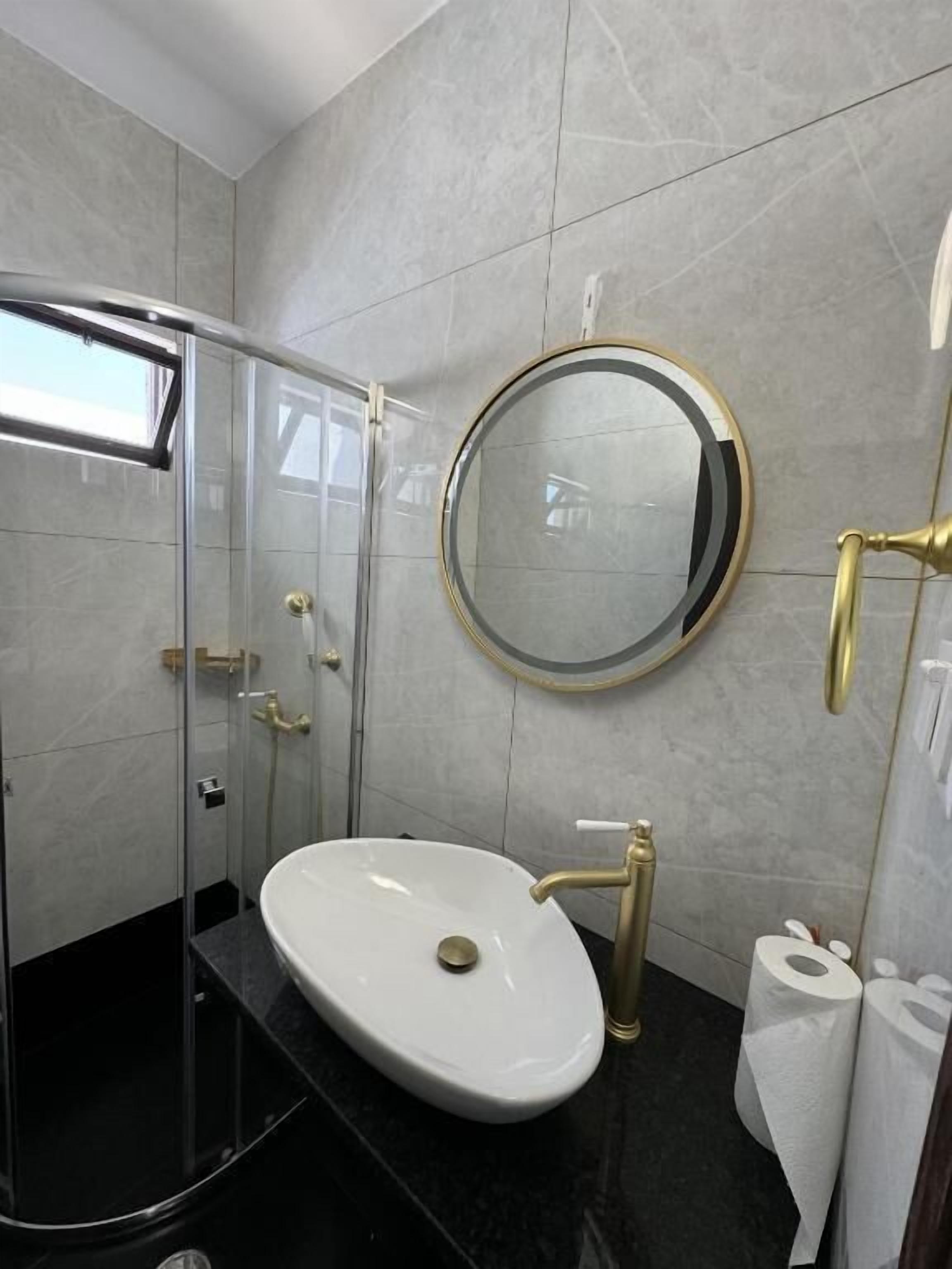 A modern bathroom with a glass shower enclosure, a round mirror, a white sink with gold fixtures, and a toilet with toilet paper.