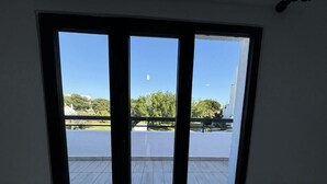 Une pièce dotée d’une grande fenêtre donnant sur un balcon, d’où l’on peut admirer des arbres et un immeuble.