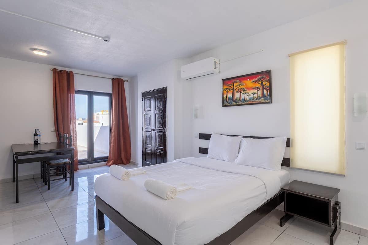 A hotel room with a bed, a desk, a chair, a painting, and an air conditioning unit.