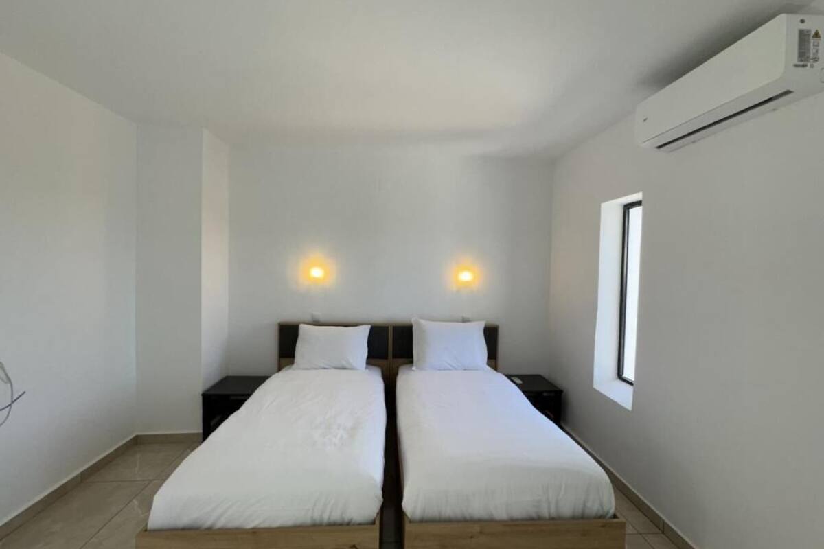 A room with two single beds, a wooden floor, a white wall, a window, and an air conditioning unit.