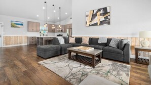 A spacious living room with a sectional sofa, a coffee table, and a rug. There is a kitchen in the background with modern appliances and pendant lighting. - Huge Vacation Home! Community Boardwalk to the Beach (Port Aransas)