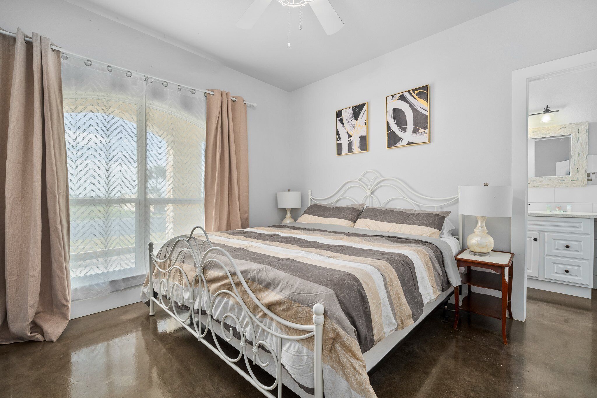 A bedroom with a bed, curtains, a window, a bedside table, a lamp, and a mirror.