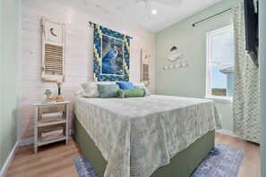 A bedroom with a bed, a nightstand, a window with curtains, and a wall with a bird painting. - St. Jo's Bungalow (Port Aransas)