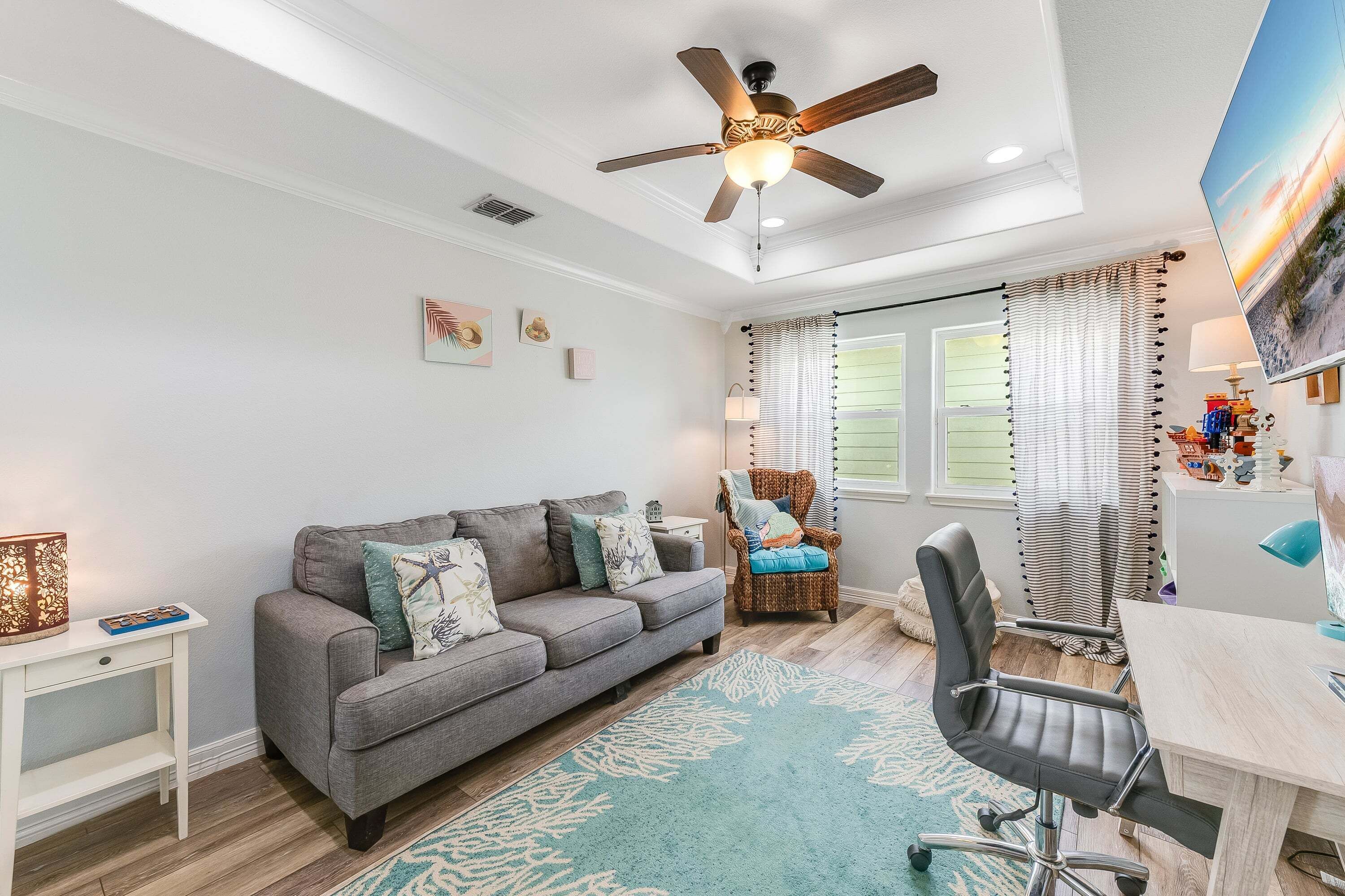 A modern living room with a grey sofa, a wicker chair, a desk, and a ceiling fan.