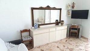 A room with a white sofa, a wooden chair, a white cabinet with decorative vases, a large mirror, and a television on a stand. - Laura's House - Aspra Sea View (Bagheria)