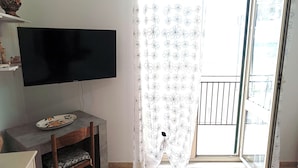 A room with a flat-screen TV on a stand, a small table with a plate, a chair, a patterned curtain, and a sliding glass door leading to an outdoor area. - Laura's House - Aspra Sea View (Bagheria)