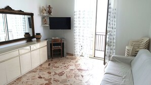 A living room with a stone floor, a white sofa, a TV on a stand, a mirror, and a window with curtains. - Laura's House - Aspra Sea View (Bagheria)