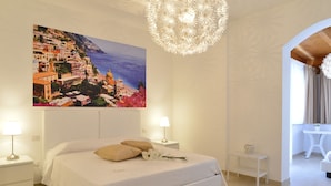 A bedroom with a large bed, two bedside tables, a nightstand, a hanging light fixture, and a painting of a coastal town. - Villa Miriam (Gragnano)