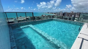 A rooftop infinity pool with clear blue water, surrounded by glass railings, offering a view of the ocean and city skyline. - Smart Stay Apto 807-Mandi Hospitalidade (Maceio)