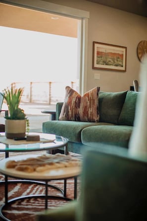 A green sofa with a patterned pillow, a glass coffee table, a potted plant, and a framed picture on the wall.