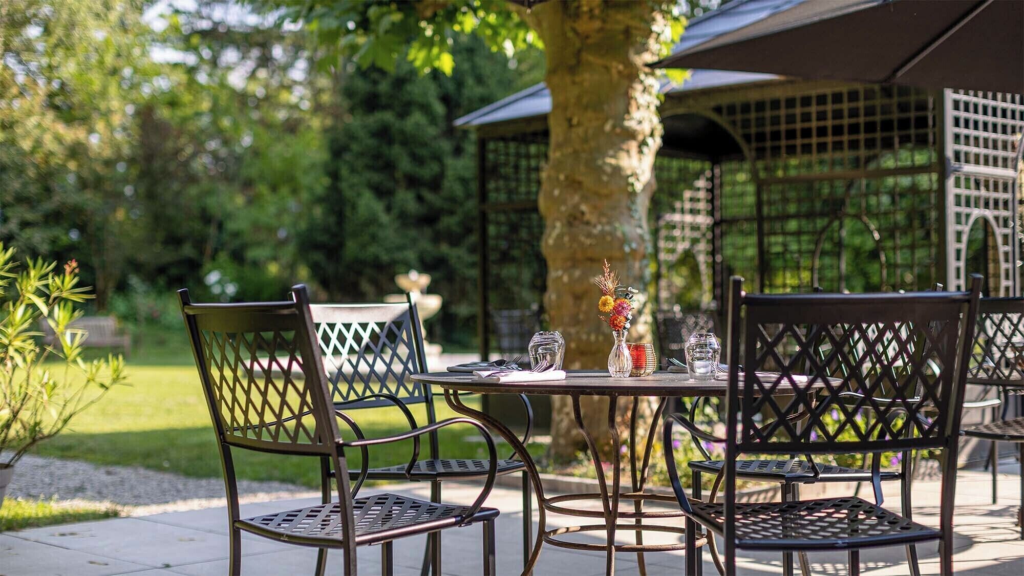 An outdoor dining area with a metal table and chairs, surrounded by greenery and a pergola structure.