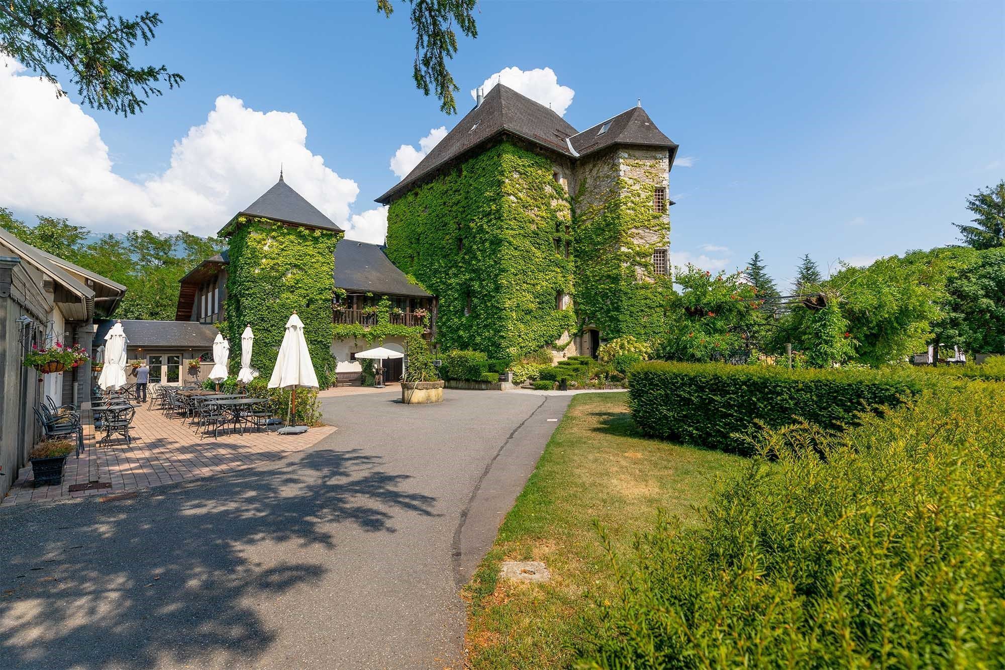 Un bâtiment traditionnel en pierre, avec des murs recouverts de lierre et une tour, entouré d’une cour pavée aménagée avec des espaces de détente en plein air et des parasols.
