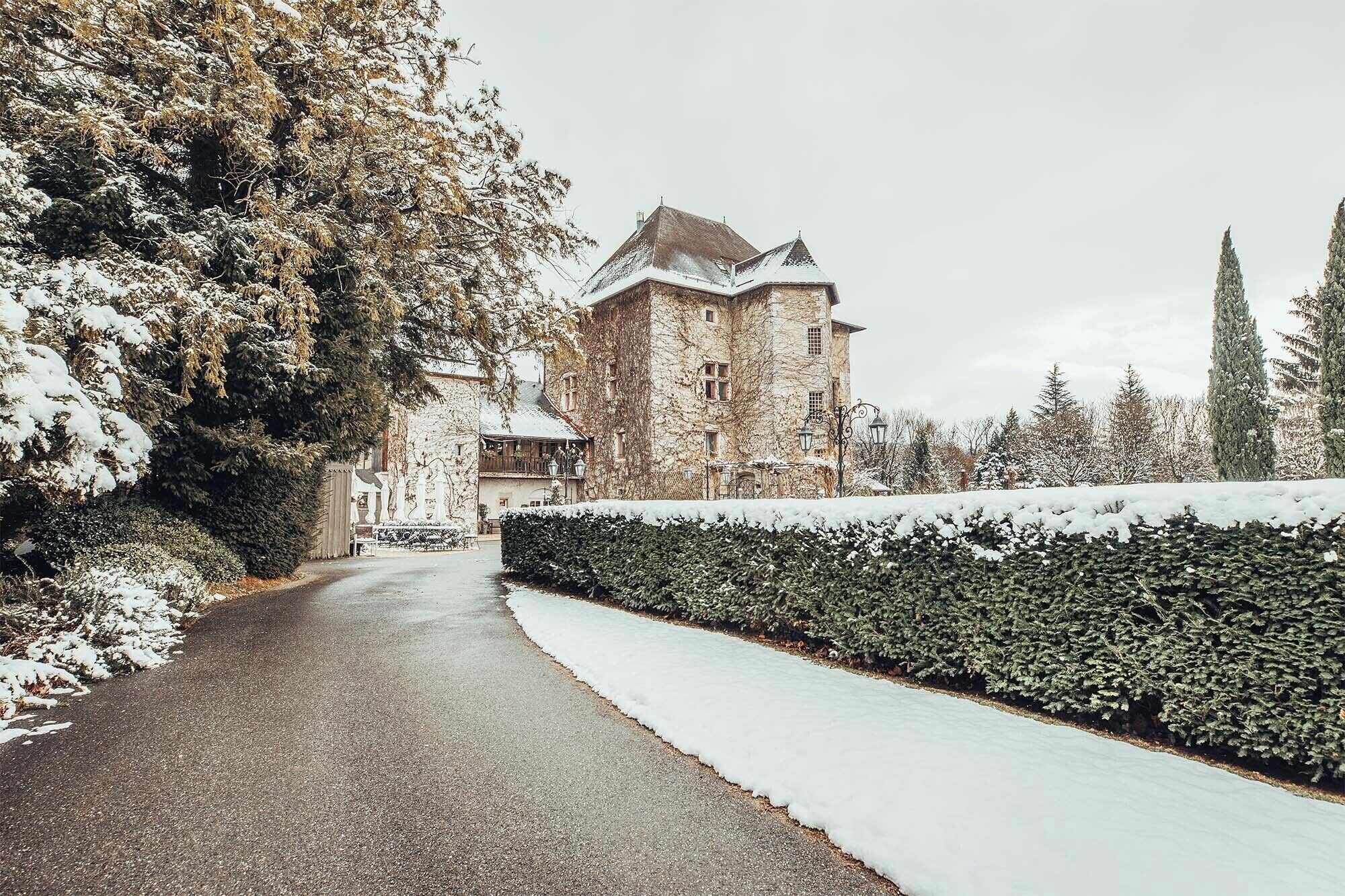 Un sentier enneigé mène à un bâtiment en pierre au style traditionnel, entouré d’arbres à feuilles persistantes et d’un arbuste taillé avec soin.