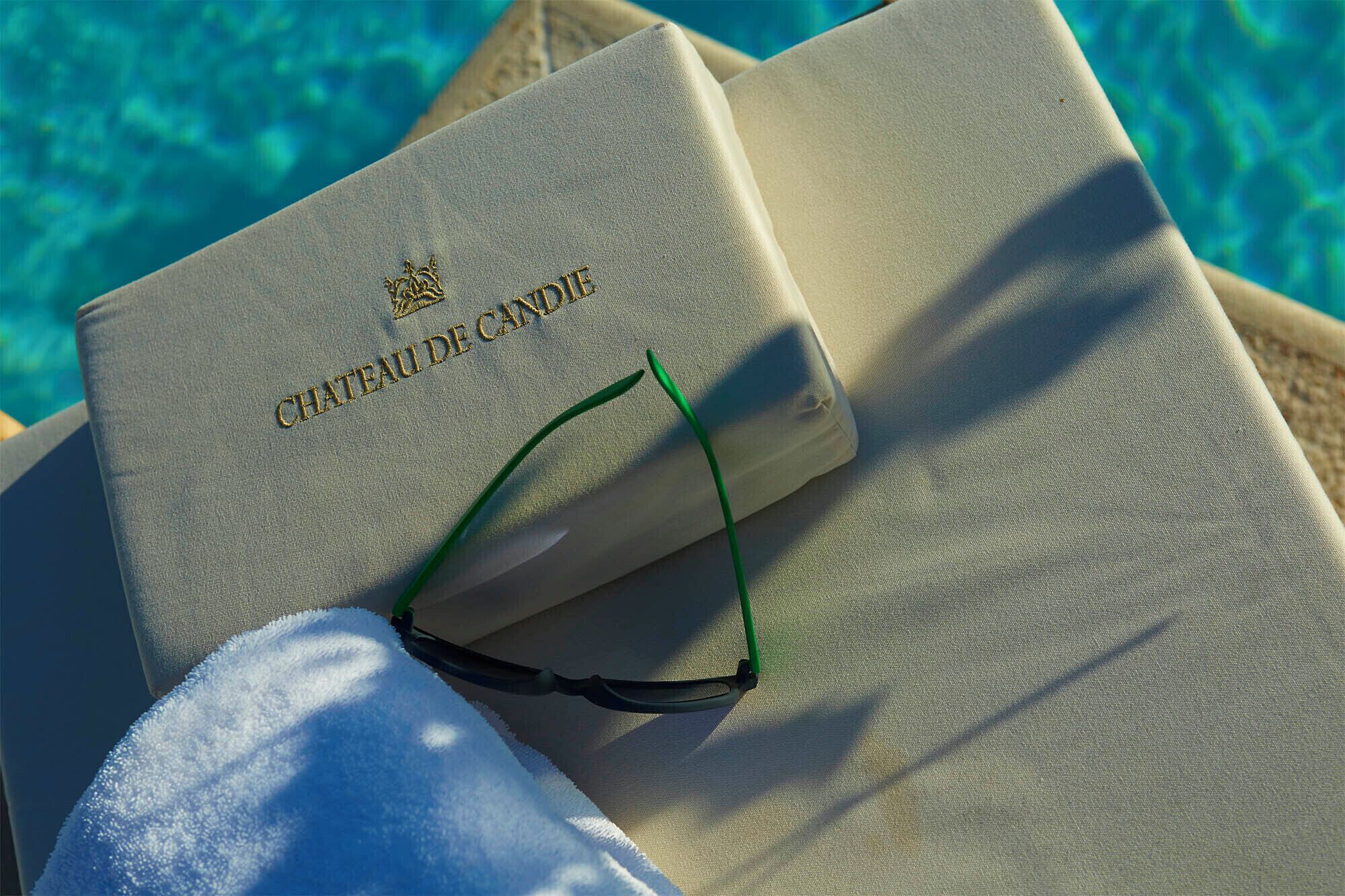 A book titled "CHATEAU DE CANDIE" with a crown emblem, placed on a lounge chair by a pool.