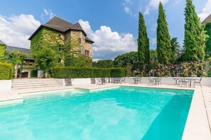 Una piscina con sillas de descanso, un edificio de piedra con hiedra y altos árboles al fondo.