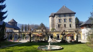 Un edificio histórico de piedra con una torre, un patio con una fuente y zonas de estar al aire libre.