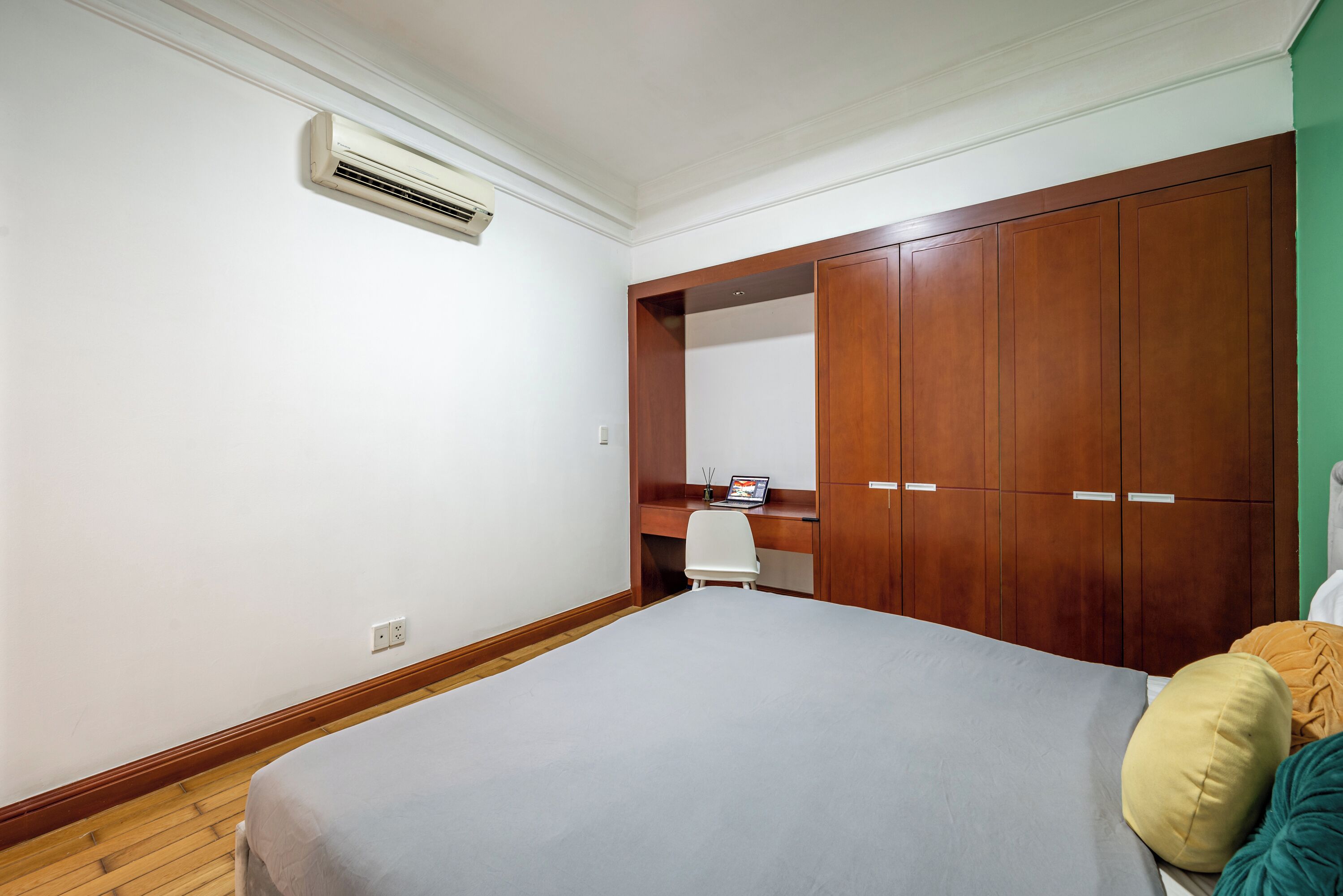 A bedroom with a bed, a wooden wardrobe, a desk with a chair, and an air conditioning unit.